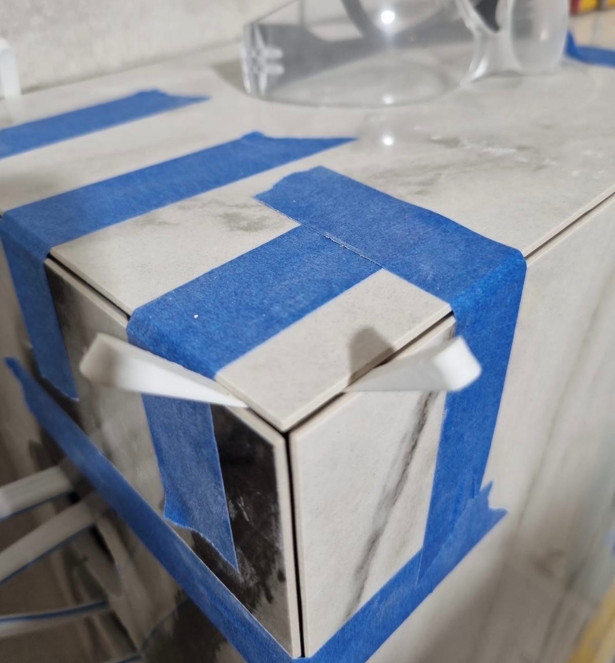 Fragment wykończenia z płytek imitujących marmur, zabezpieczonych niebieską taśmą malarską. Widoczne okulary ochronne w tle. Detal narożnika.