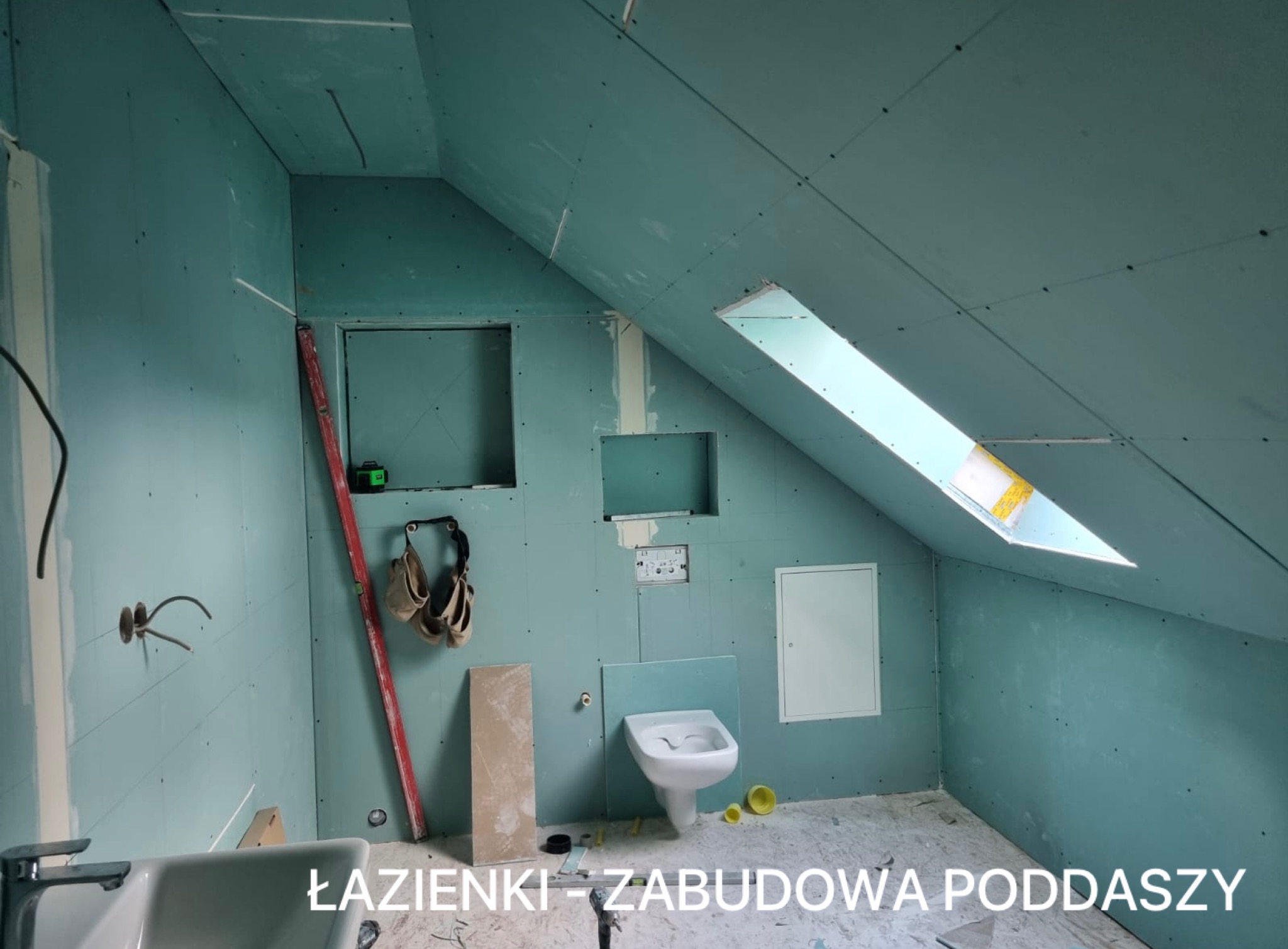 Poddasze w trakcie adaptacji na łazienkę: ściany obłożone płytami gips-karton, widoczne instalacje, okno dachowe i fragment umywalki. Etap prac wykończeniowych.