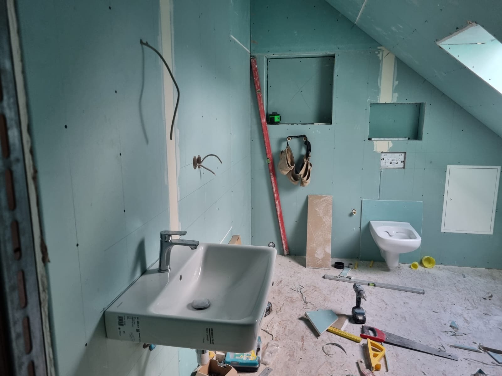 Łazienka na poddaszu w trakcie prac: montaż umywalki, płyty gipsowe, instalacja elektryczna. Widoczne narzędzia i poziomica. Skosy sufitu i okno dachowe.