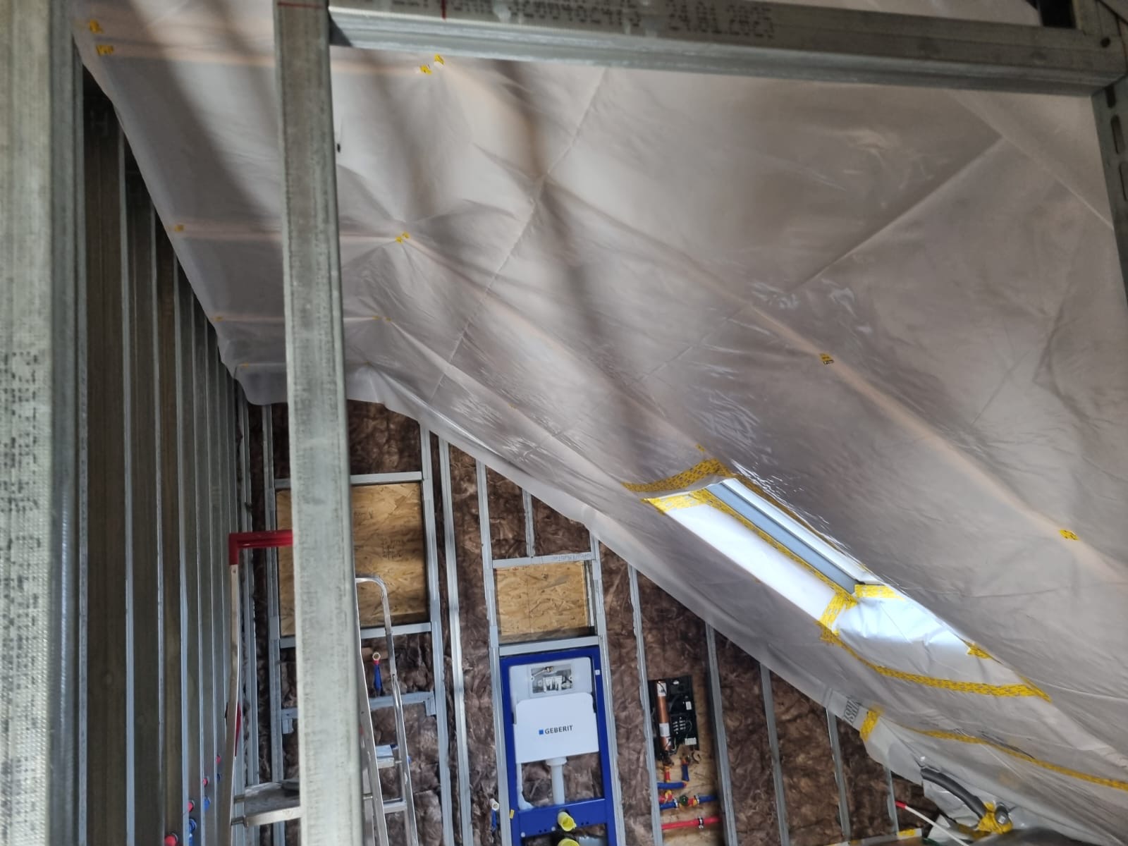 Skośne poddasze w trakcie adaptacji: metalowy stelaż, izolacja z wełny mineralnej, folia paroizolacyjna, instalacja sanitarna Geberit. Widoczna drabina i okno dachowe.