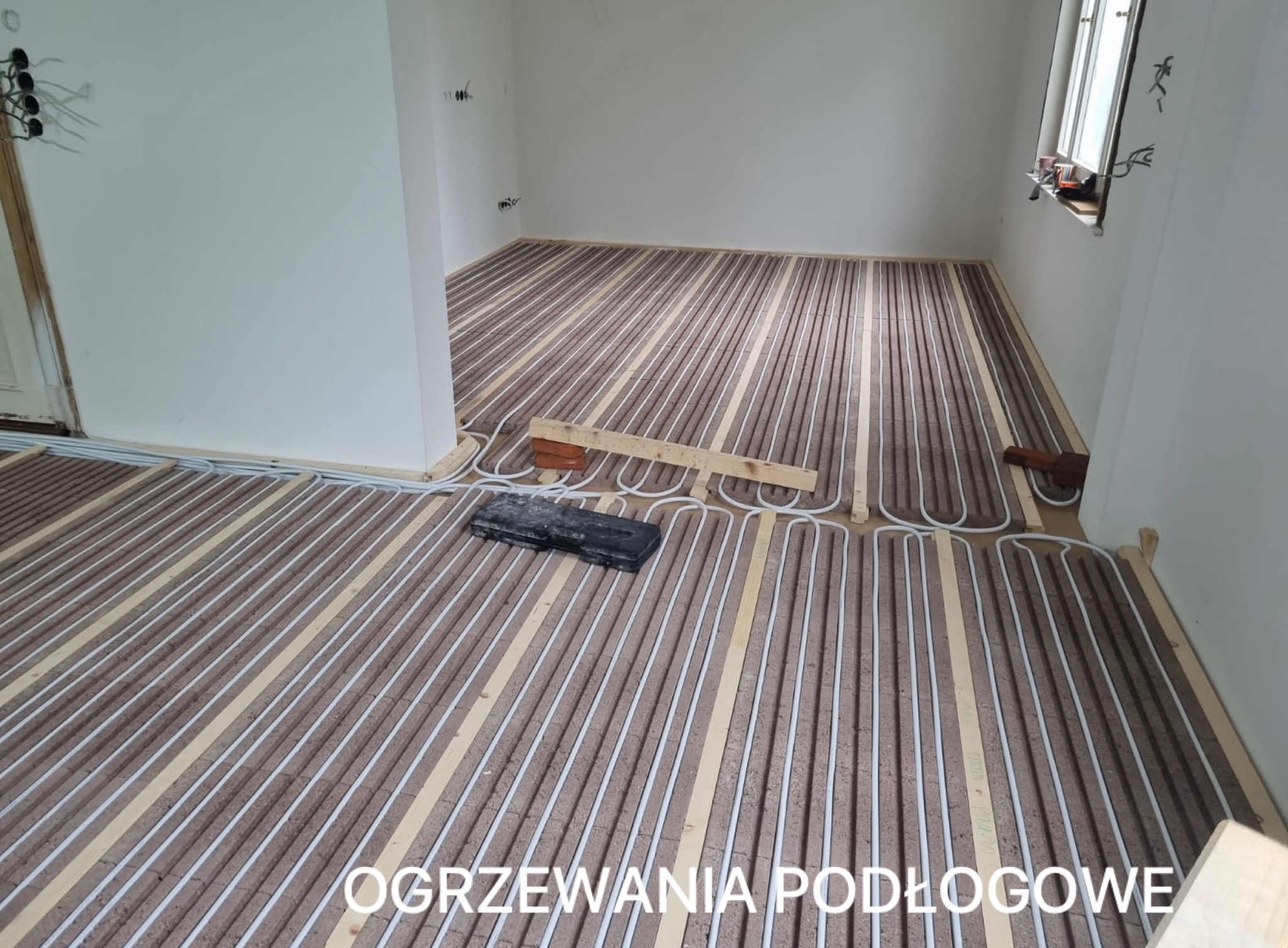 Białe rurki ogrzewania podłogowego ułożone na brązowej macie w jasnym pomieszczeniu. Widoczne narzędzia i elementy montażowe. Etap instalacji przed wylaniem posadzki.