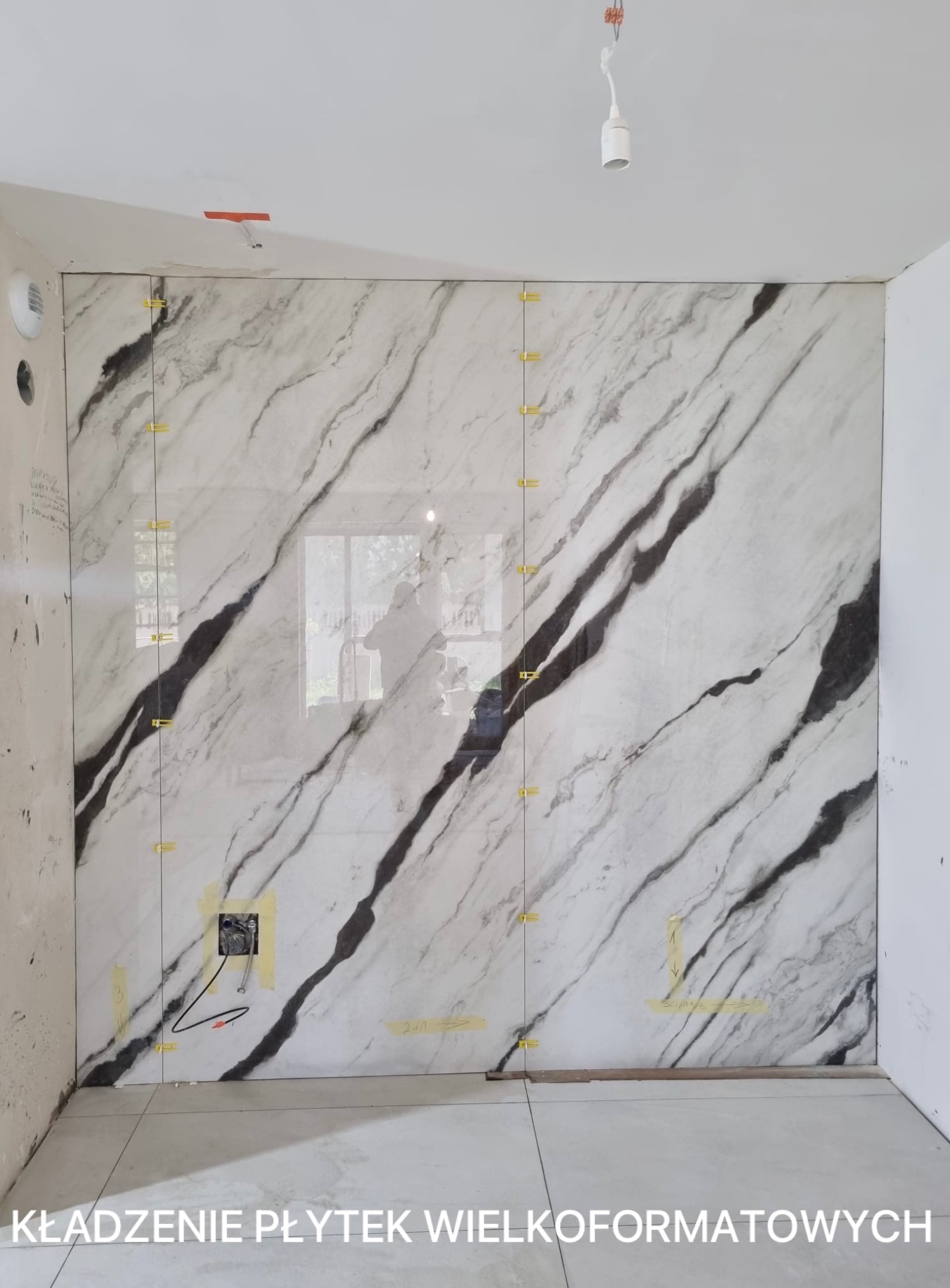 Montaż wielkoformatowych płytek imitujących marmur na ścianie w łazience, z widocznymi żółtymi krzyżykami dystansowymi i puszką elektryczną. Etap prac wykończeniowych.