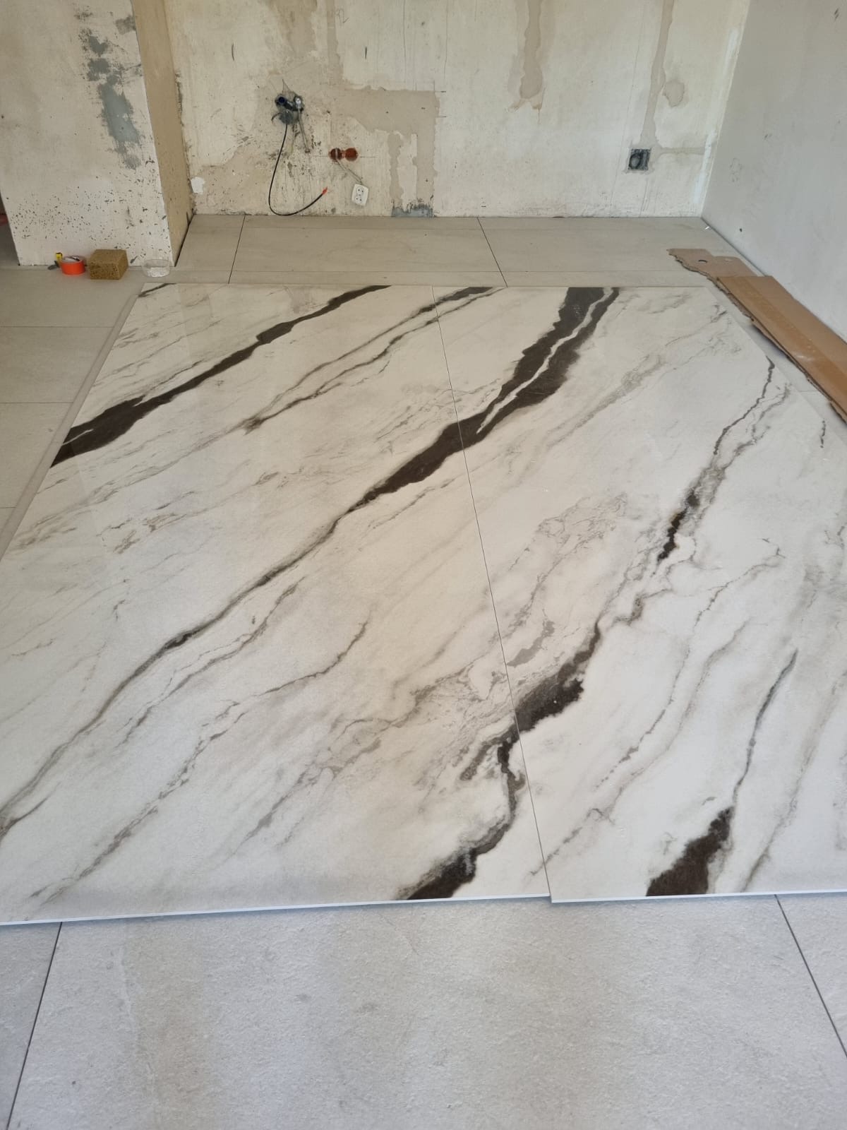 Ułożone, marmurowe płytki podłogowe z ciemnymi żyłami, kontrastujące z surowym tłem ściany w trakcie prac. Widoczne detale instalacji elektrycznej.