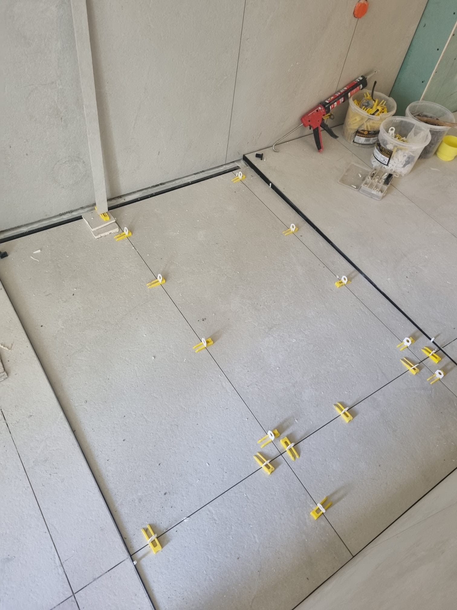 Układanie jasnych płytek podłogowych z użyciem żółtych klipsów poziomujących i czarnych listew. Widoczne narzędzia i pojemniki z akcesoriami.