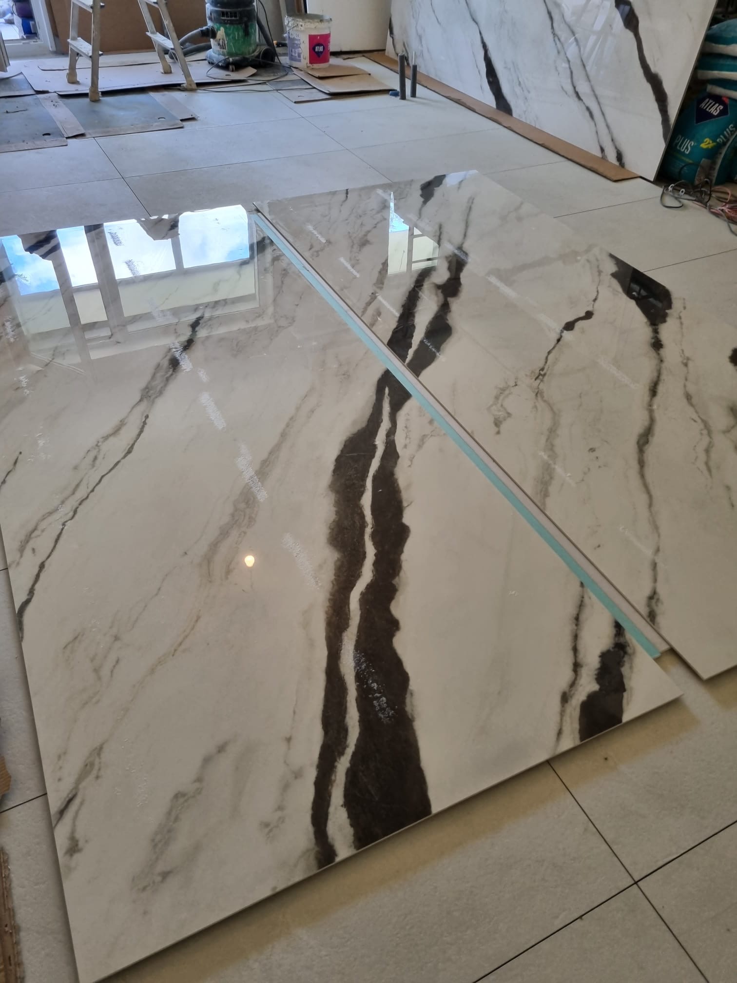 Błyszczące, wielkoformatowe płytki imitujące marmur z wyraźnym, ciemnym żyłowaniem, ułożone na podłodze. Widoczne narzędzia i materiały budowlane w tle.