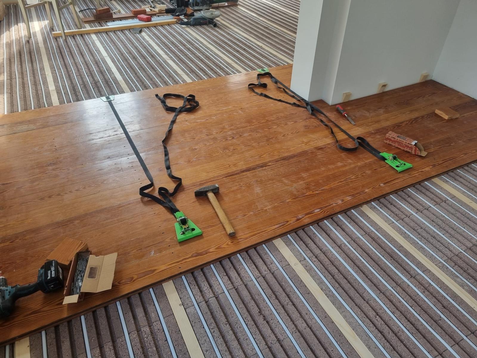 Układanie drewnianej podłogi na ogrzewaniu podłogowym. Widoczne narzędzia: młotek, wkrętarka, napinacz. Częściowo ułożona podłoga z desek o ciepłym odcieniu brązu.