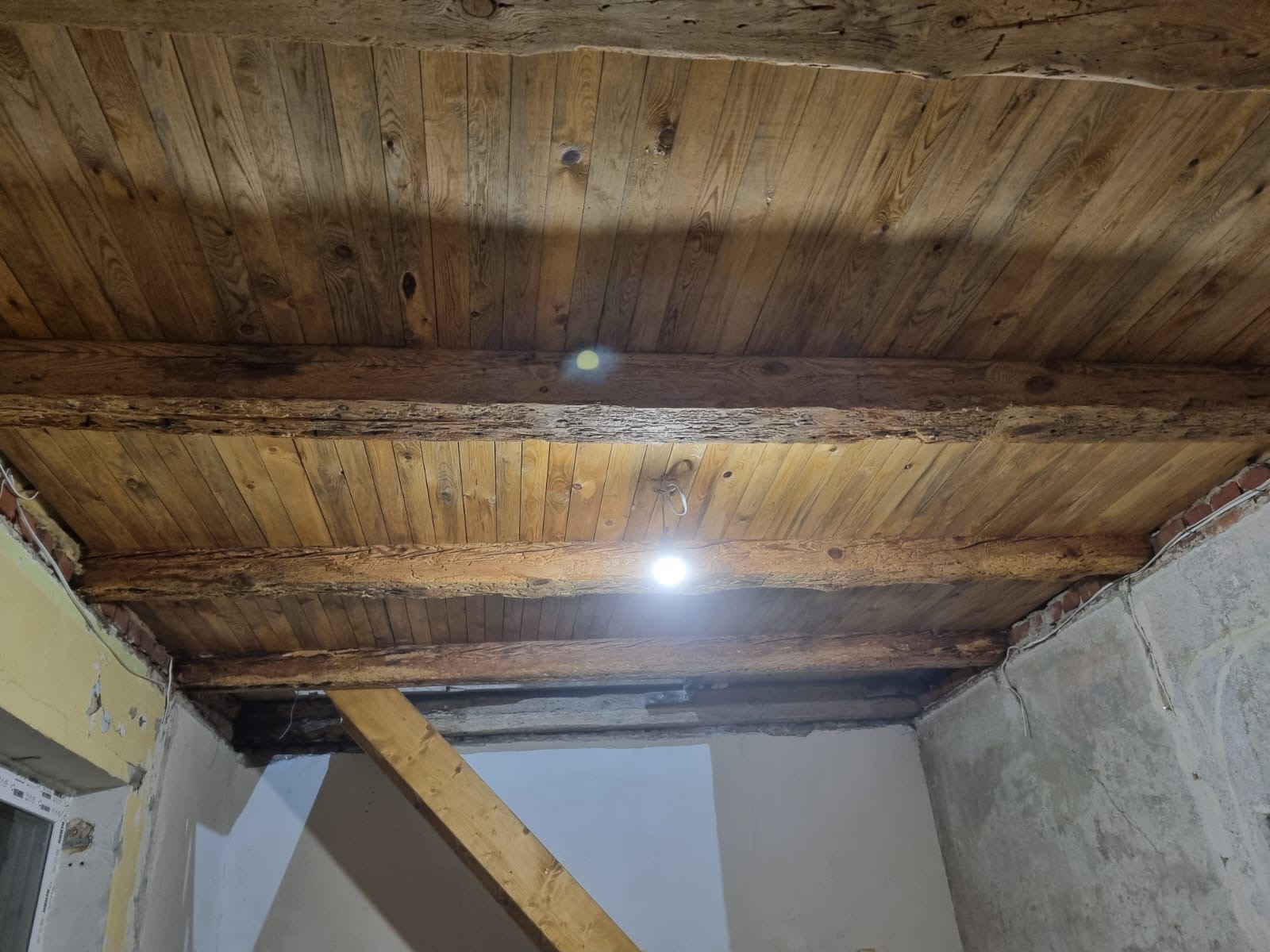 Drewniany sufit z belkami w trakcie prac remontowych. Widoczne fragmenty ścian w stanie surowym i instalacja elektryczna. Oświetlenie punktowe podkreśla strukturę drewna.