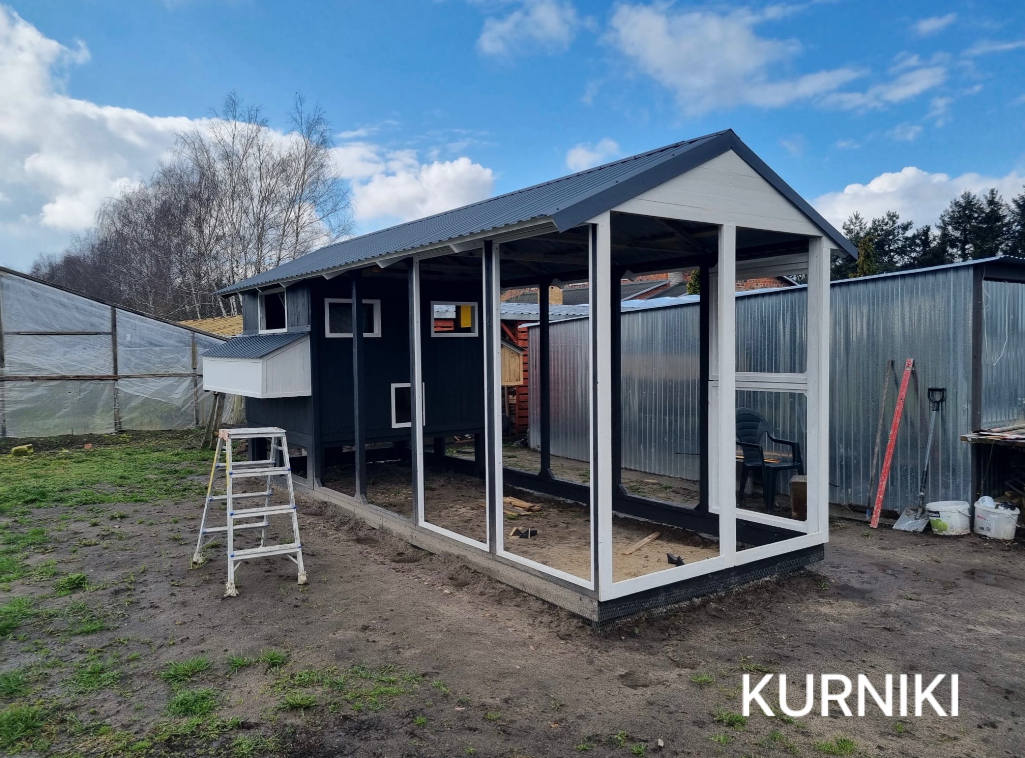 Nowoczesny, czarno-biały kurnik z metalowym dachem, częściowo otwarty, z drabiną z boku. W tle szklarnia i budynek gospodarczy, błękitne niebo.