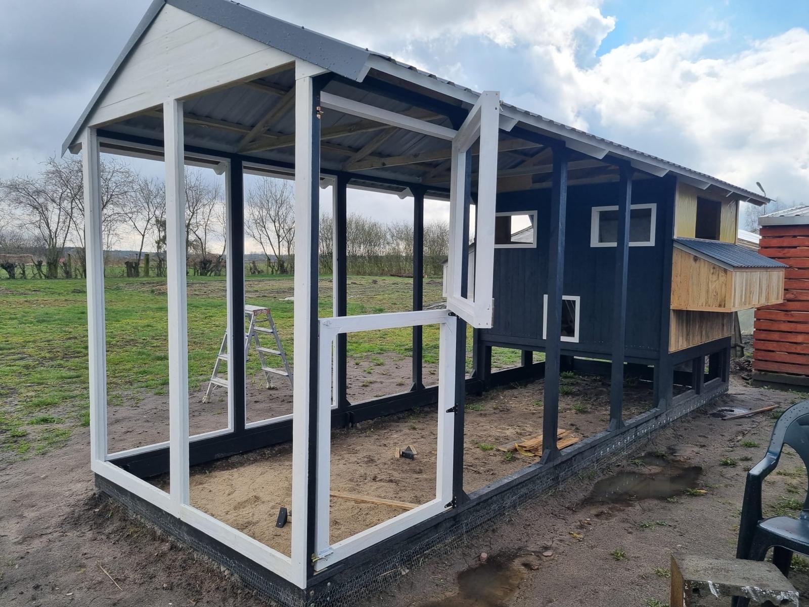 Częściowo ukończona, czarno-biała konstrukcja drewniana z otwartymi ramami okiennymi i drabiną w tle, na zewnątrz, w trakcie budowy. Widoczne elementy wykończeniowe.