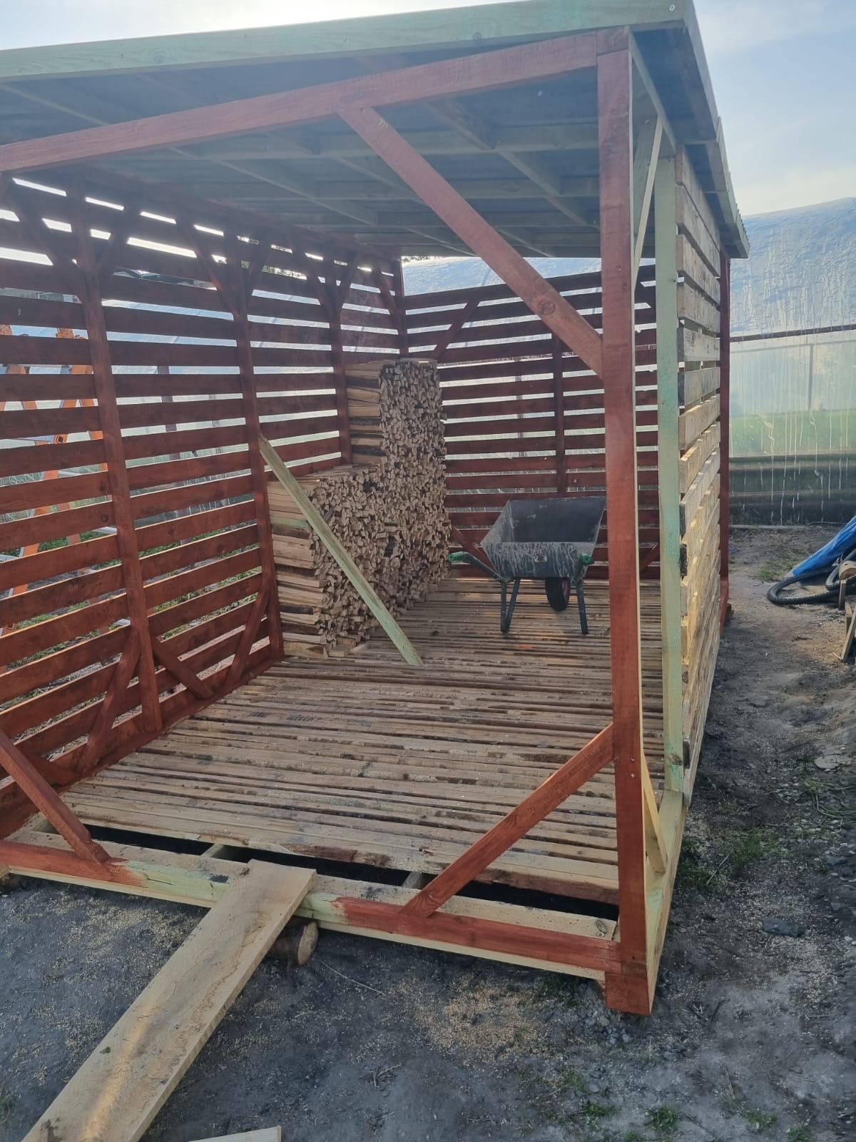Drewniana konstrukcja z palet w trakcie budowy, widoczny szkielet i podłoga, w tle sterta drewna i taczka. Konstrukcja częściowo pomalowana na brązowo.