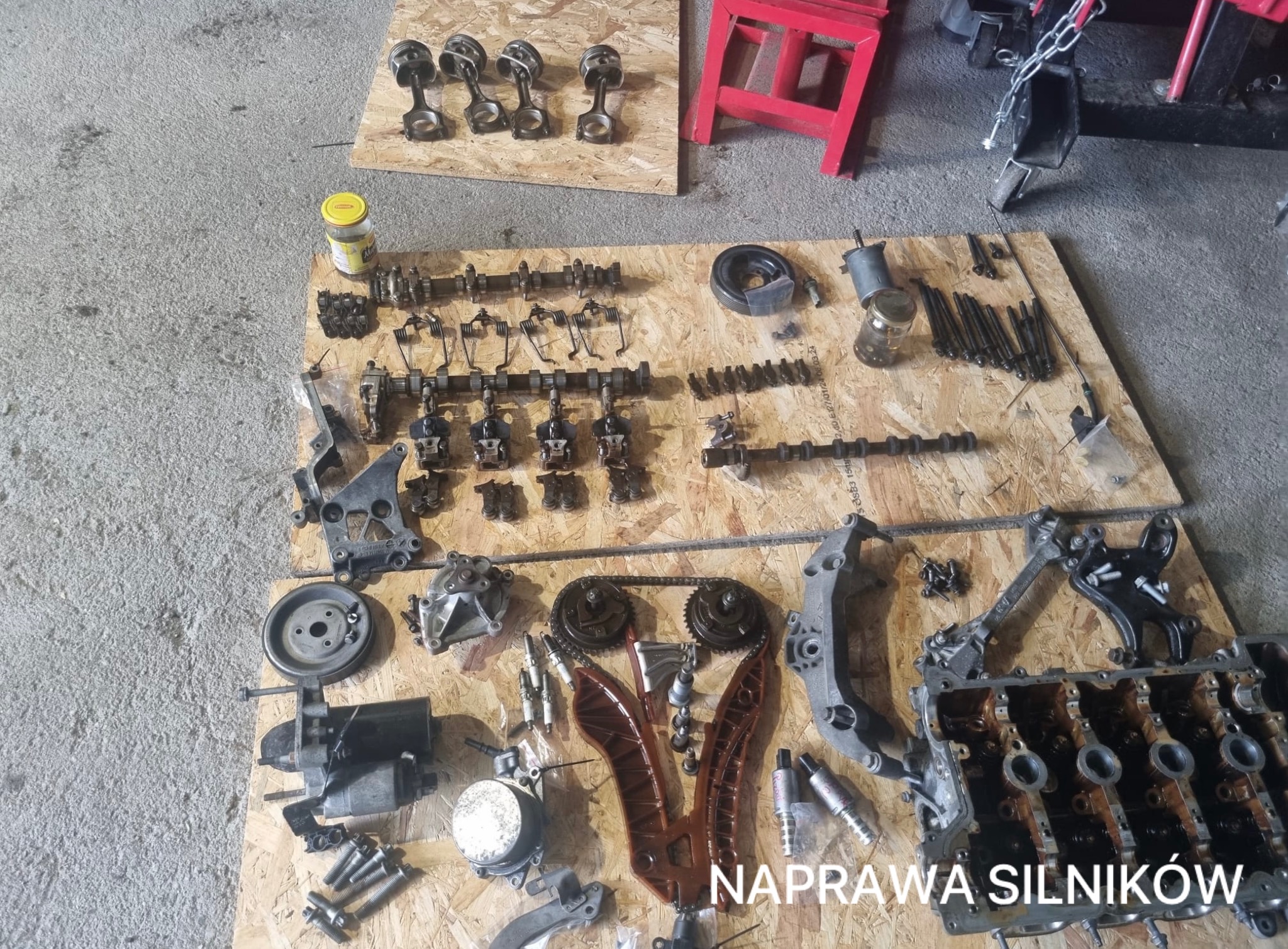 Rozłożony silnik samochodowy na drewnianej płycie. Widoczne tłoki, wałki rozrządu, łańcuch rozrządu i inne elementy. Naprawa silników.