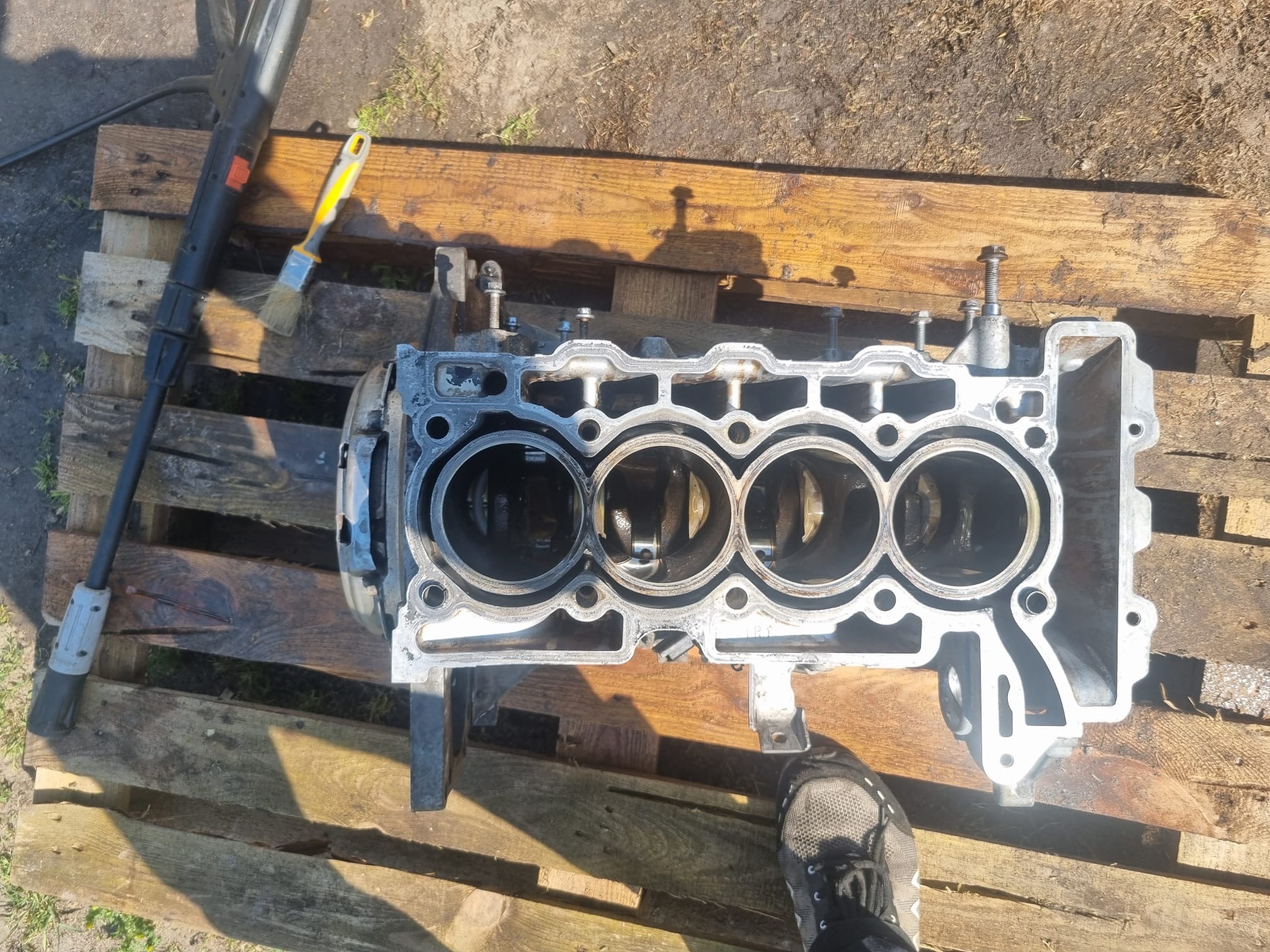 Czyszczenie bloku silnika spalinowego na drewnianej palecie, widoczna myjka ciśnieniowa i pędzel. Blok czterocylindrowy, z widocznymi tłokami. Noga w bucie w prawym dolnym rogu.