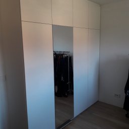 JUST.LUXURY BARBARA GROBLICKA, MONIKA GÓRNIAK-SEROWIK S.C. - Biała szafa wnękowa z lustrem, widok z boku, na podłodze dokumenty. Minimalistyczny design, jasne wnętrze, odbicie w lustrze ukazuje ubrania na wieszakach.