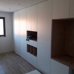 JUST.LUXURY BARBARA GROBLICKA, MONIKA GÓRNIAK-SEROWIK S.C. - Biała szafa wnękowa z otwartymi półkami i miejscem na ubrania, widoczna na tle jasnego wnętrza z drewnianą podłogą i oknem. Minimalistyczny design.