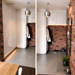 Forma Meble - KUCHNIE - SZAFY - GARDEROBY - Biała szafa wnękowa z oświetleniem LED w przedpokoju z ceglaną ścianą. Wiszące ubrania i metalowa lampa kulista dodają nowoczesnego charakteru.