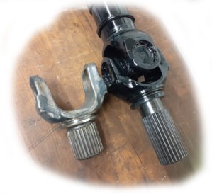 Dwa wały napędowe, jeden z widocznymi frezami, drugi czarny, błyszczący, leżące na drewnianej powierzchni. Detal naprawy lub wymiany elementu układu napędowego.