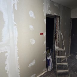 Fine Space Remonty - Ściana z płyt kartonowo-gipsowych w trakcie szpachlowania, widoczne łączenia i miejsca po wkrętach. W tle drabina i worki z materiałami budowlanymi.
