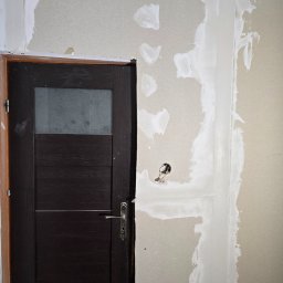 Fine Space Remonty - Montaż płyty gipsowej z widocznymi śladami szpachlowania przy futrynie drzwiowej z ciemnego drewna i szklanym panelem. Widoczne kable elektryczne w ścianie.
