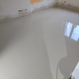 Fine Space Remonty - Wylana, gładka posadzka samopoziomująca w jasnym odcieniu szarości, odbijająca światło okna. Ściany częściowo przygotowane do malowania, widoczne gniazdka elektryczne.