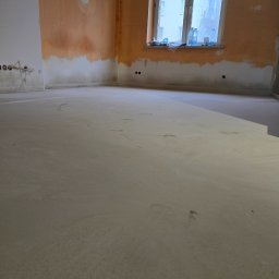 Fine Space Remonty - Wnętrze w trakcie prac: wylewka podłogowa, ściany częściowo przygotowane do malowania, widoczne instalacje elektryczne. Okno z widokiem na budynek.