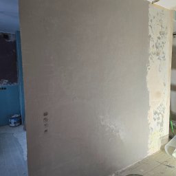 Fine Space Remonty - Ściana po szpachlowaniu z widoczną siatką wzmacniającą, fragment ściany przed obróbką, krzesło i wiadro farby w tle. Etap prac wykończeniowych w pomieszczeniu.