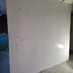 Fine Space Remonty - Ściana po szpachlowaniu, widoczne ślady po wałku. Fragment sufitu z widocznym ubytkiem. W tle fragment pomieszczenia z puszkami elektrycznymi i farbą.