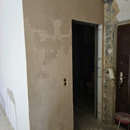 Fine Space Remonty - Ściana po szpachlowaniu, widoczne nierówności i faktura nałożonej gładzi. Obok fragment ściany przygotowanej do malowania, drzwi wejściowe i fragment toalety w głębi.