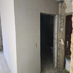 Fine Space Remonty - Ściana po szpachlowaniu z widoczną fakturą, fragment ściany pomalowany na biało, drzwi do łazienki w tle, widoczne ślady remontu, kubeł z farbą na stołku.