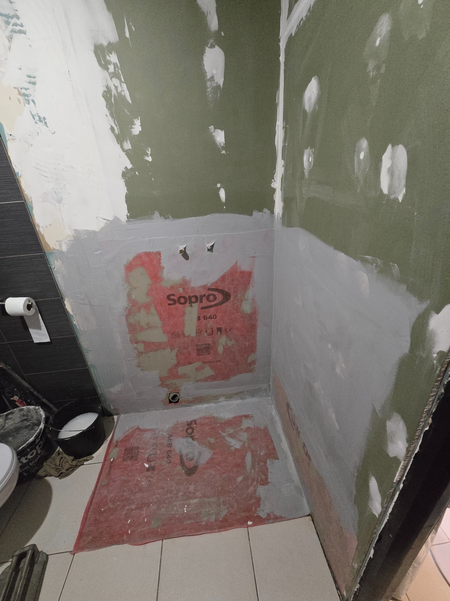 Przygotowanie ścian w łazience pod płytki: gruntowanie, hydroizolacja Sopro, częściowo położona gładź. Widoczne narzędzia i wiaderka. Etap prac wykończeniowych.