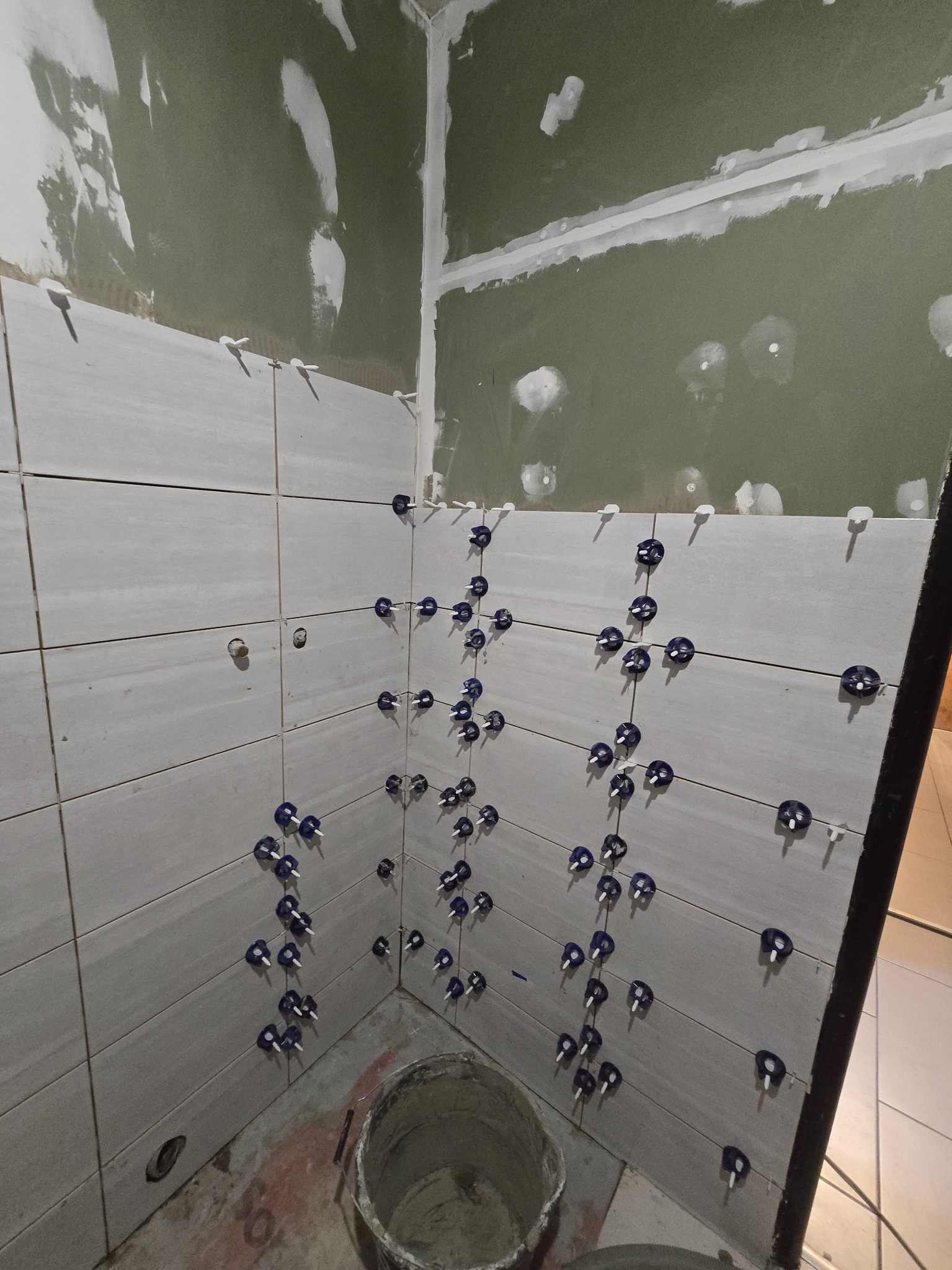 Ściana w trakcie układania płytek, z użyciem niebieskich klipsów poziomujących. Widoczne surowe płyty karton-gips na górze ściany i wiadro na podłodze.