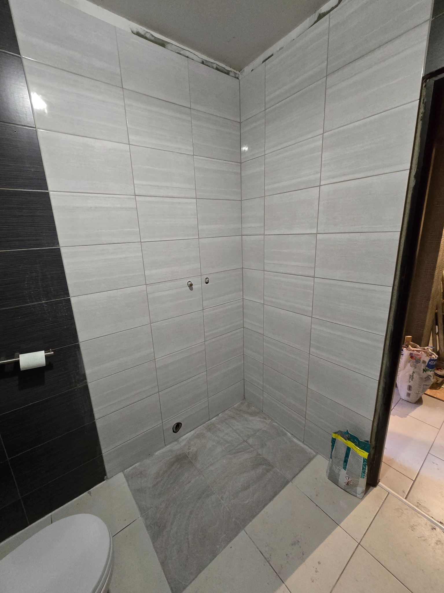 Wykończona łazienka z szaro-białymi płytkami imitującymi kamień, czarny akcent na ścianie, widoczne przyłącza wodne i toaleta. Torba z klejem do płytek w rogu.