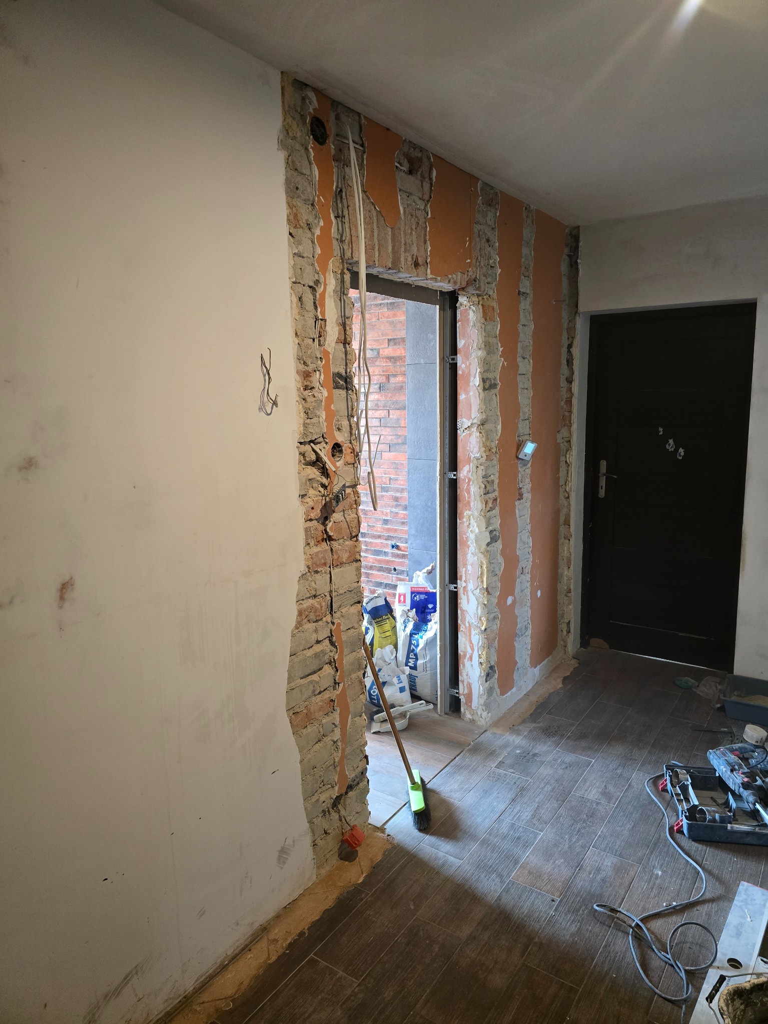 Demontaż ściany działowej z odsłoniętą cegłą i instalacją elektryczną. Widoczne narzędzia i worki z zaprawą. Podłoga wyłożona płytkami imitującymi drewno.