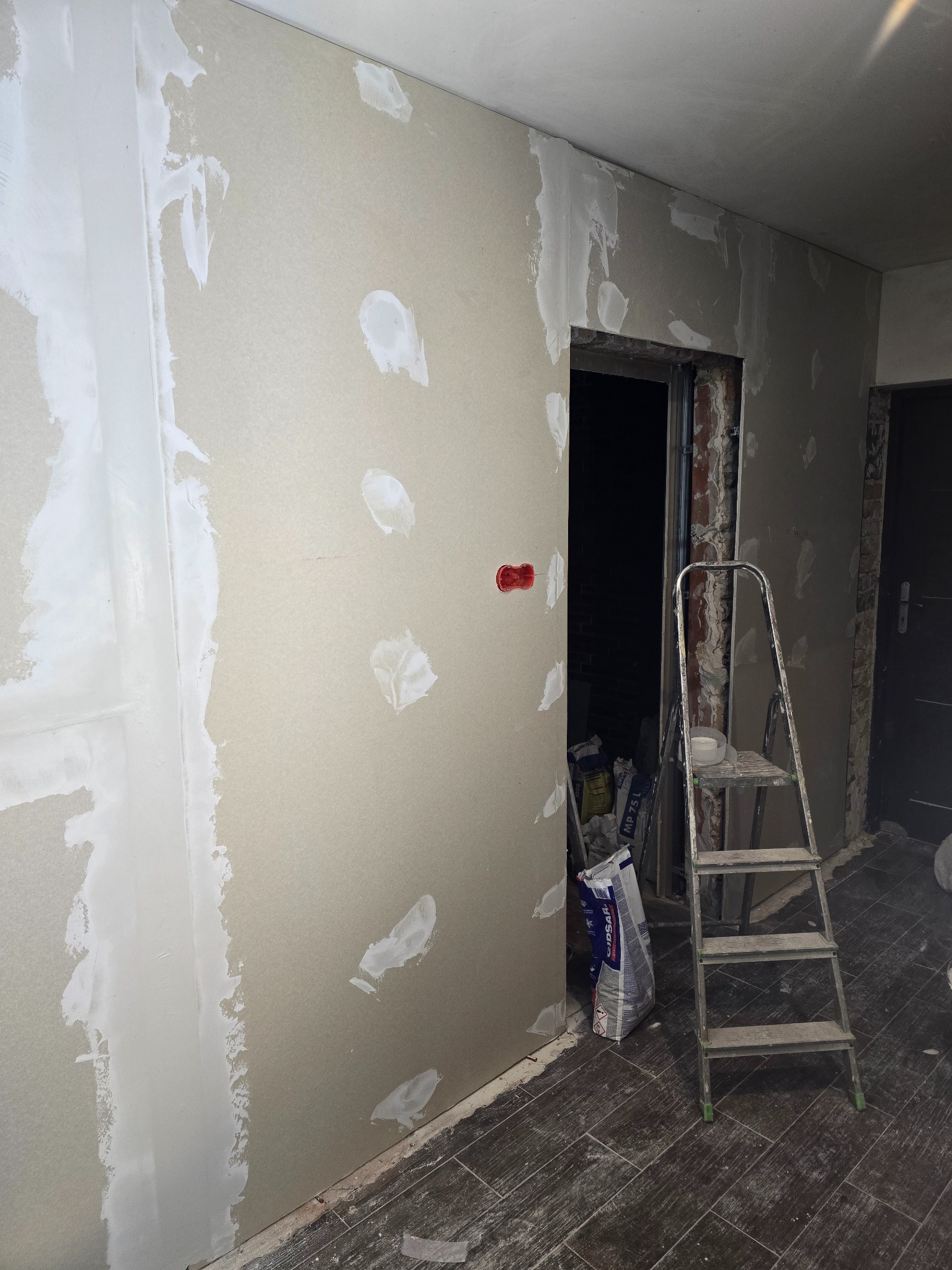 Ściana z płyt kartonowo-gipsowych w trakcie szpachlowania, widoczne łączenia i miejsca po wkrętach. W tle drabina i worki z materiałami budowlanymi.