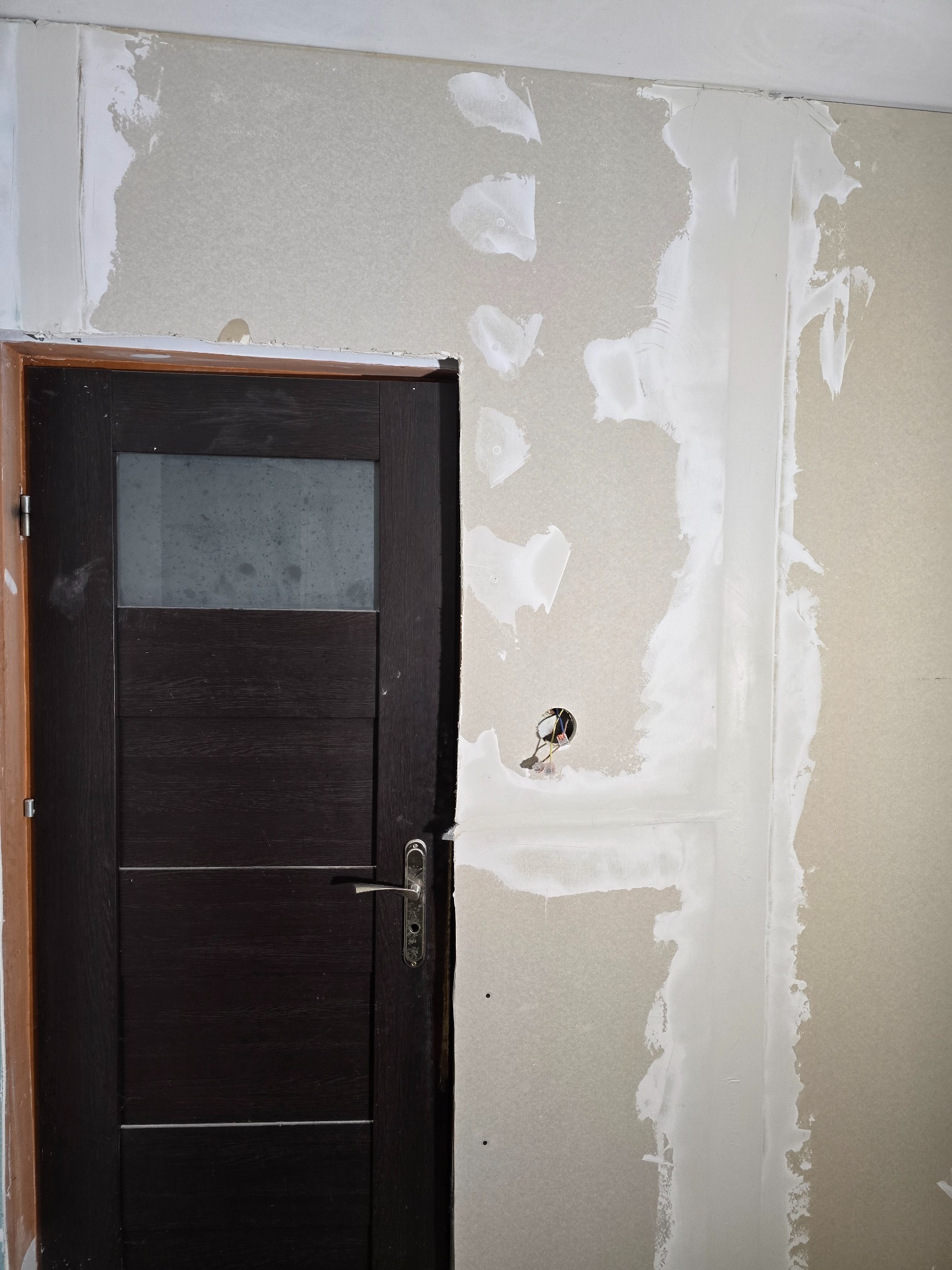 Montaż płyty gipsowej z widocznymi śladami szpachlowania przy futrynie drzwiowej z ciemnego drewna i szklanym panelem. Widoczne kable elektryczne w ścianie.