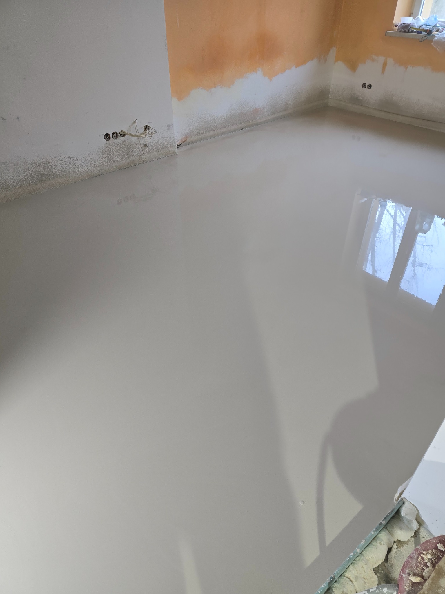 Wylana, gładka posadzka samopoziomująca w jasnym odcieniu szarości, odbijająca światło okna. Ściany częściowo przygotowane do malowania, widoczne gniazdka elektryczne.