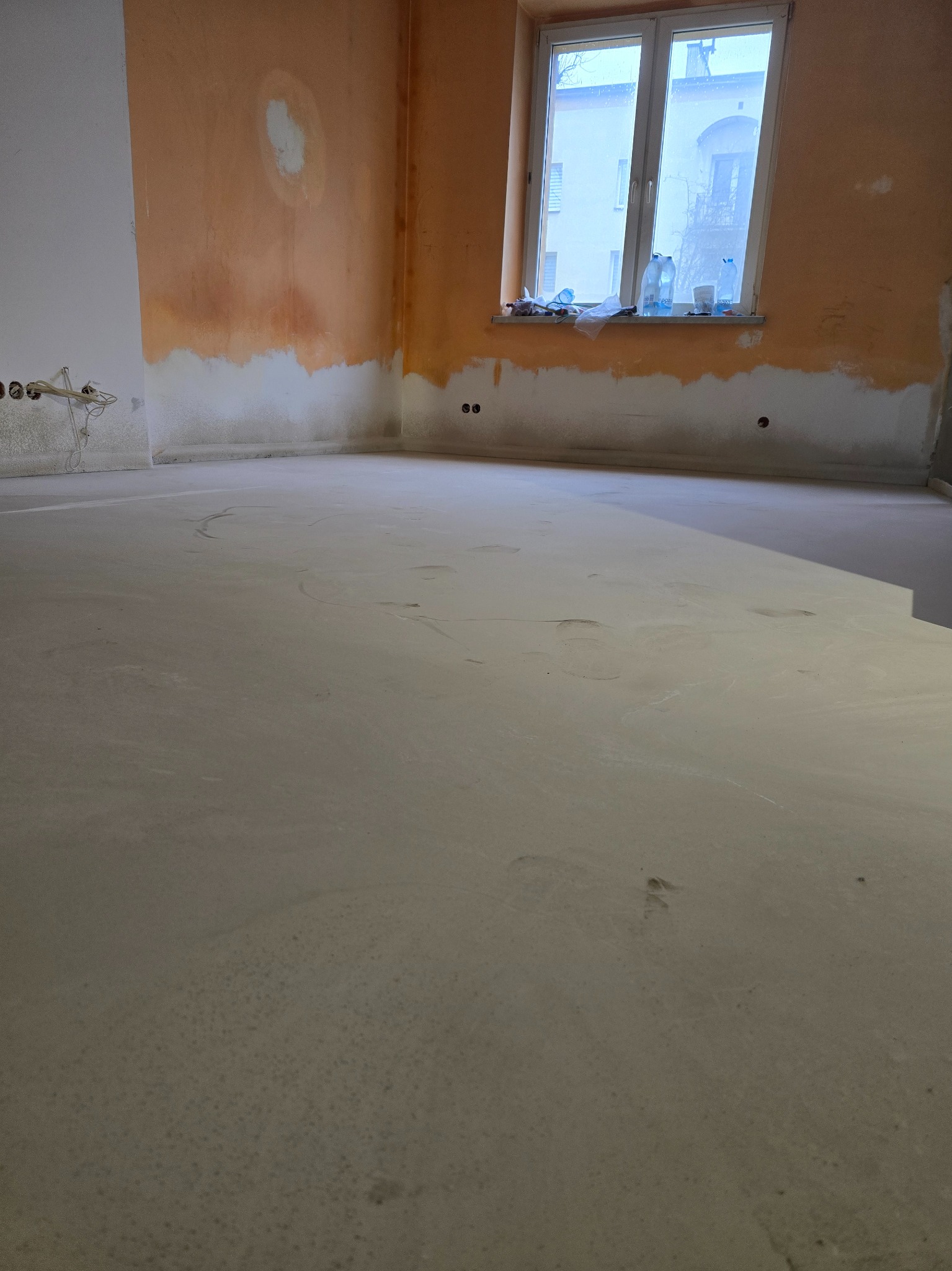 Wnętrze w trakcie prac: wylewka podłogowa, ściany częściowo przygotowane do malowania, widoczne instalacje elektryczne. Okno z widokiem na budynek.