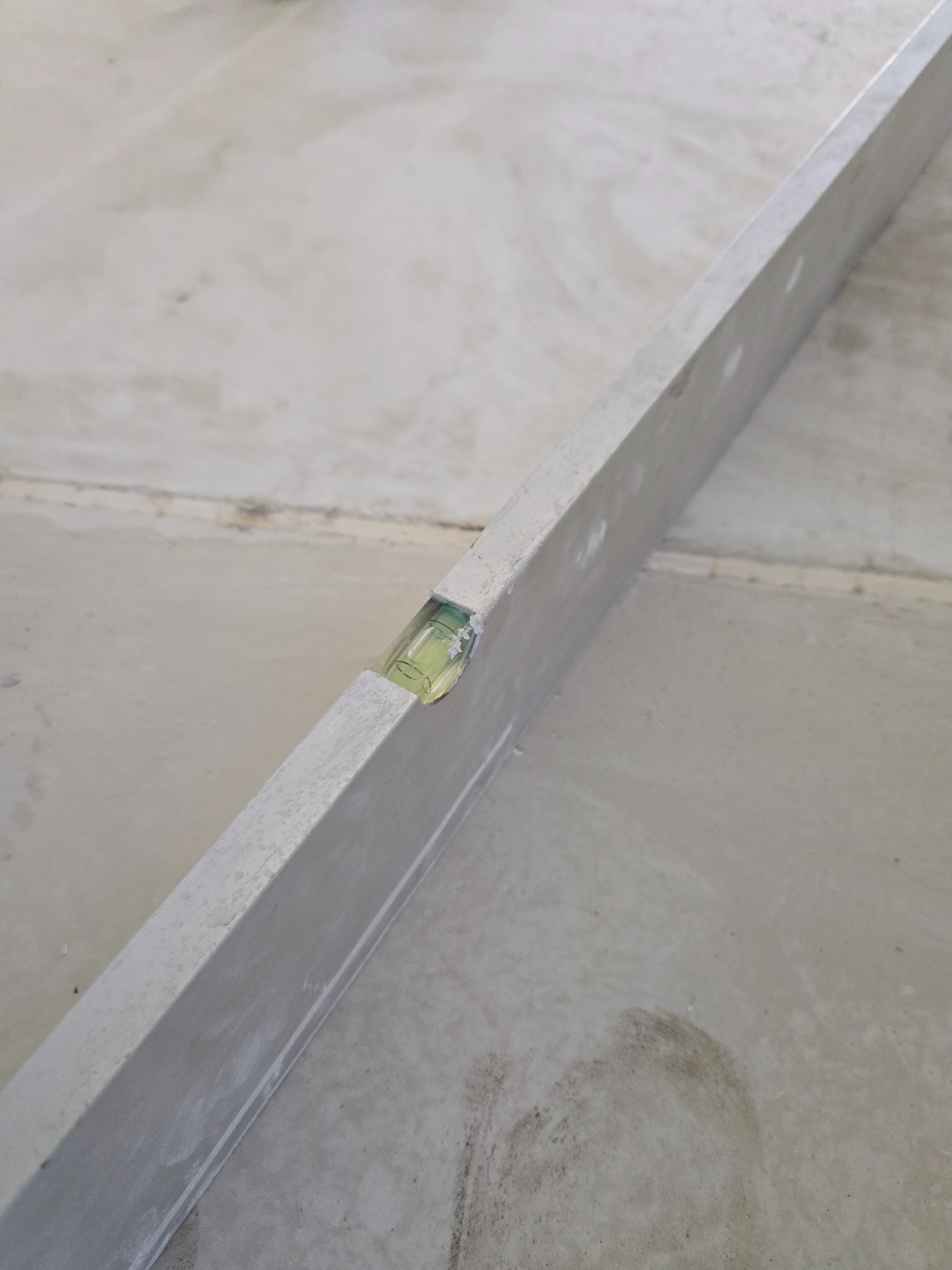 Poziomica aluminiowa na szarej, betonowej posadzce. Widoczne łączenia płyt posadzkowych. Narzędzie ustawione ukośnie, sprawdzające równość powierzchni.
