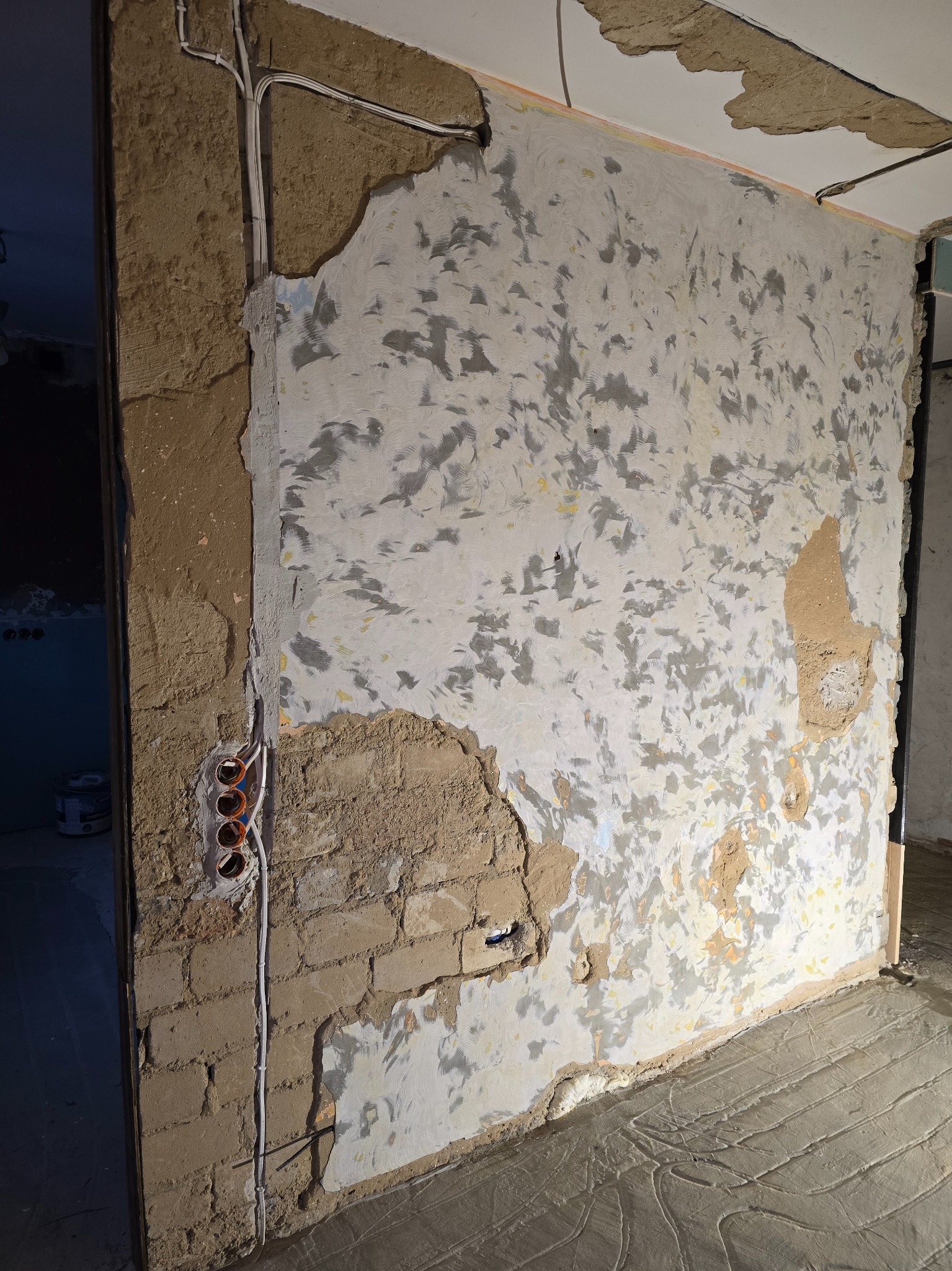 Ściana w trakcie prac przygotowawczych: częściowo otynkowana, z widocznymi fragmentami cegieł i instalacją elektryczną. Podłoga z wylewką. Widoczne ślady po usuniętych warstwach.