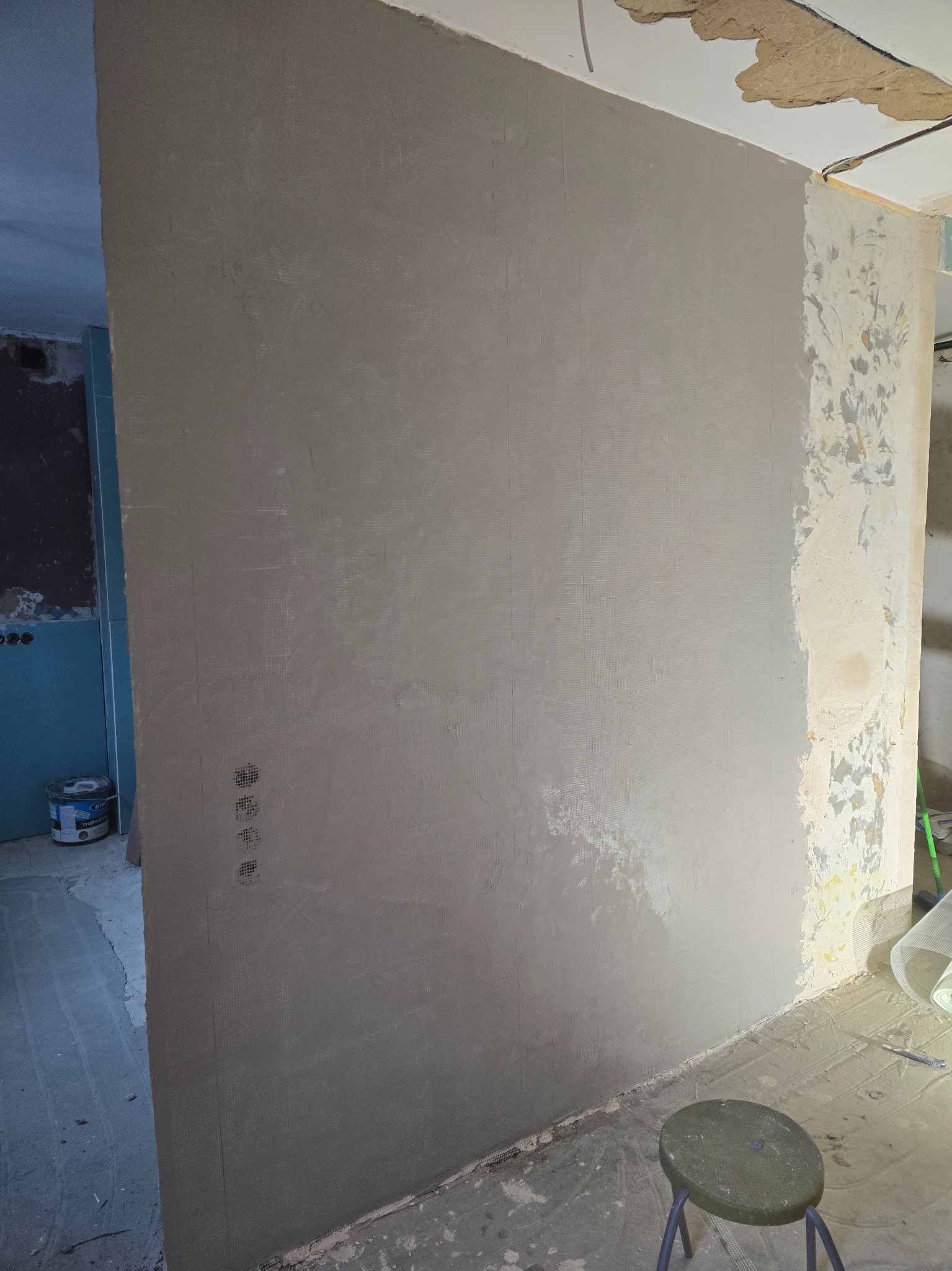 Ściana po szpachlowaniu z widoczną siatką wzmacniającą, fragment ściany przed obróbką, krzesło i wiadro farby w tle. Etap prac wykończeniowych w pomieszczeniu.