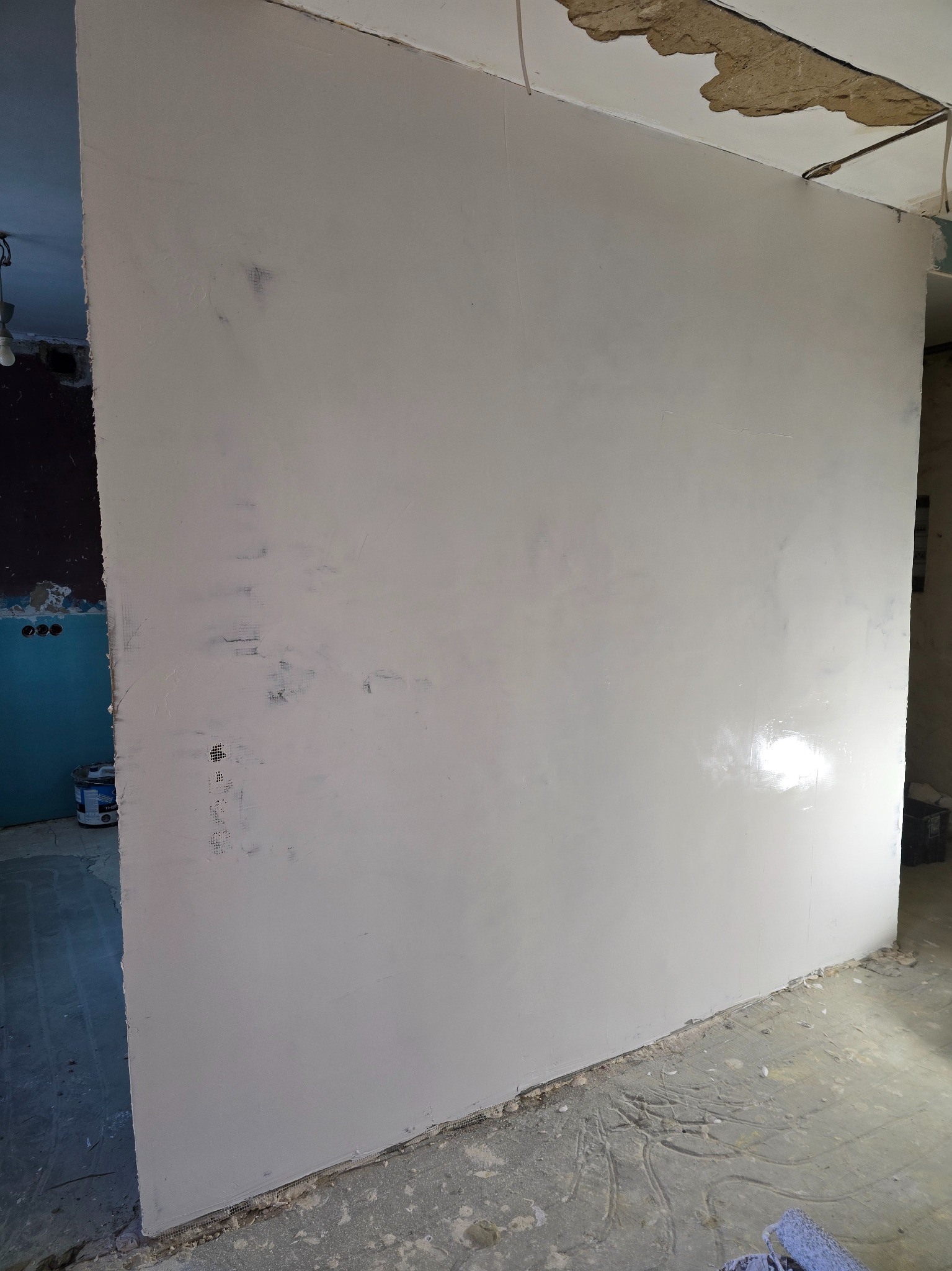 Ściana po szpachlowaniu, widoczne ślady po wałku. Fragment sufitu z widocznym ubytkiem. W tle fragment pomieszczenia z puszkami elektrycznymi i farbą.