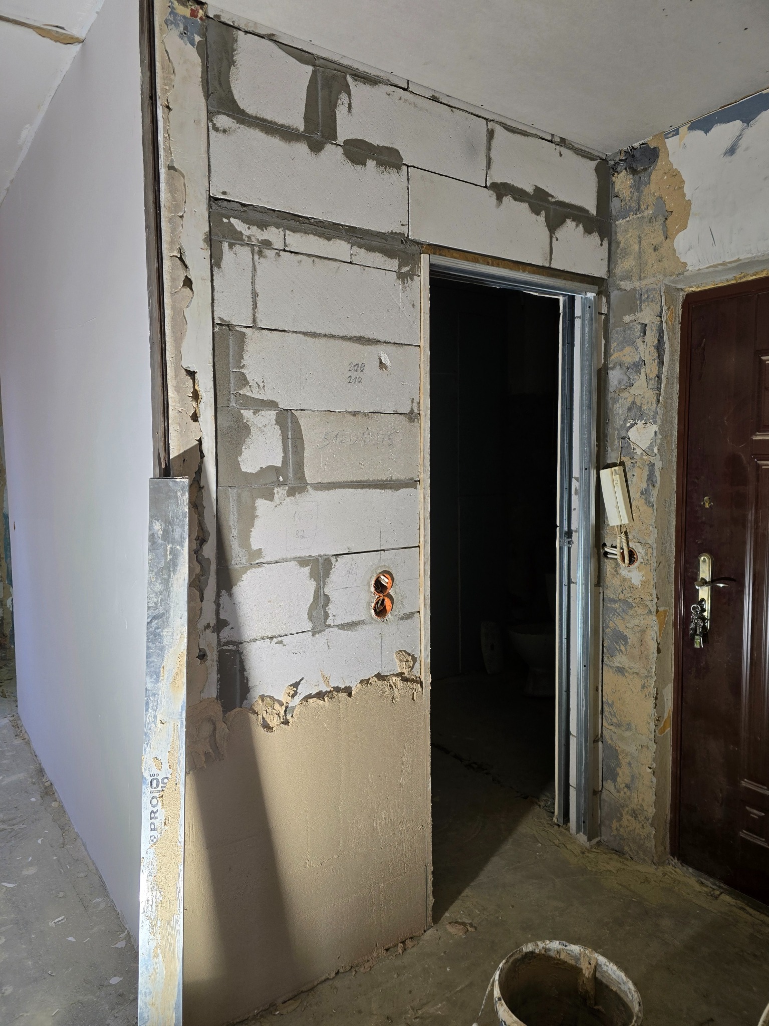 Ściana z bloków, częściowo otynkowana, z widocznymi instalacjami elektrycznymi w Zabrzu. W tle widoczne drzwi i fragment pomieszczenia w trakcie prac.