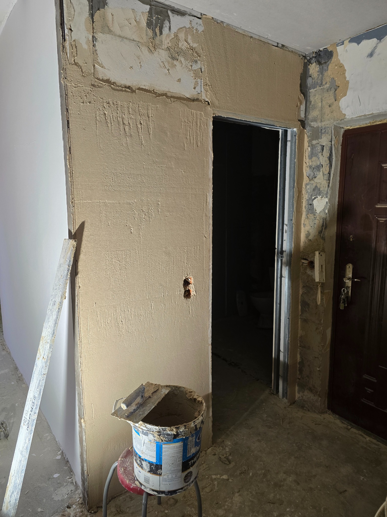 Ściana w trakcie szpachlowania z widocznym wiadrem i narzędziami, fragment płyty karton-gips i drzwi wejściowe. Widoczne ślady wcześniejszych prac remontowych.