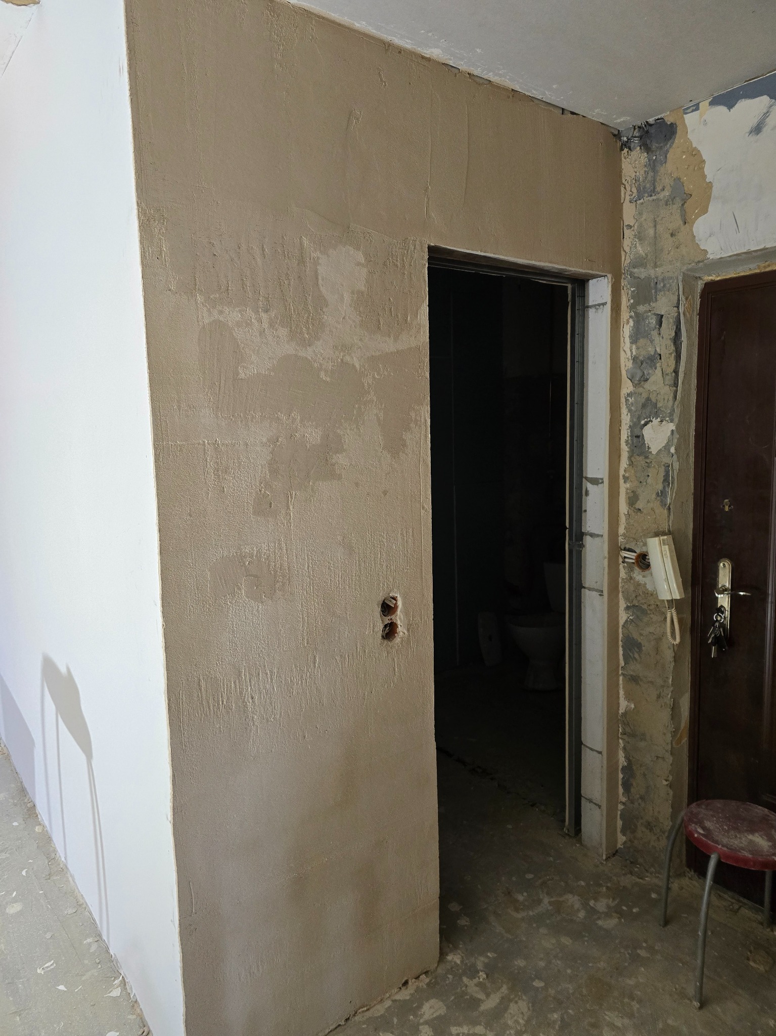 Ściana po szpachlowaniu, widoczne nierówności i faktura nałożonej gładzi. Obok fragment ściany przygotowanej do malowania, drzwi wejściowe i fragment toalety w głębi.