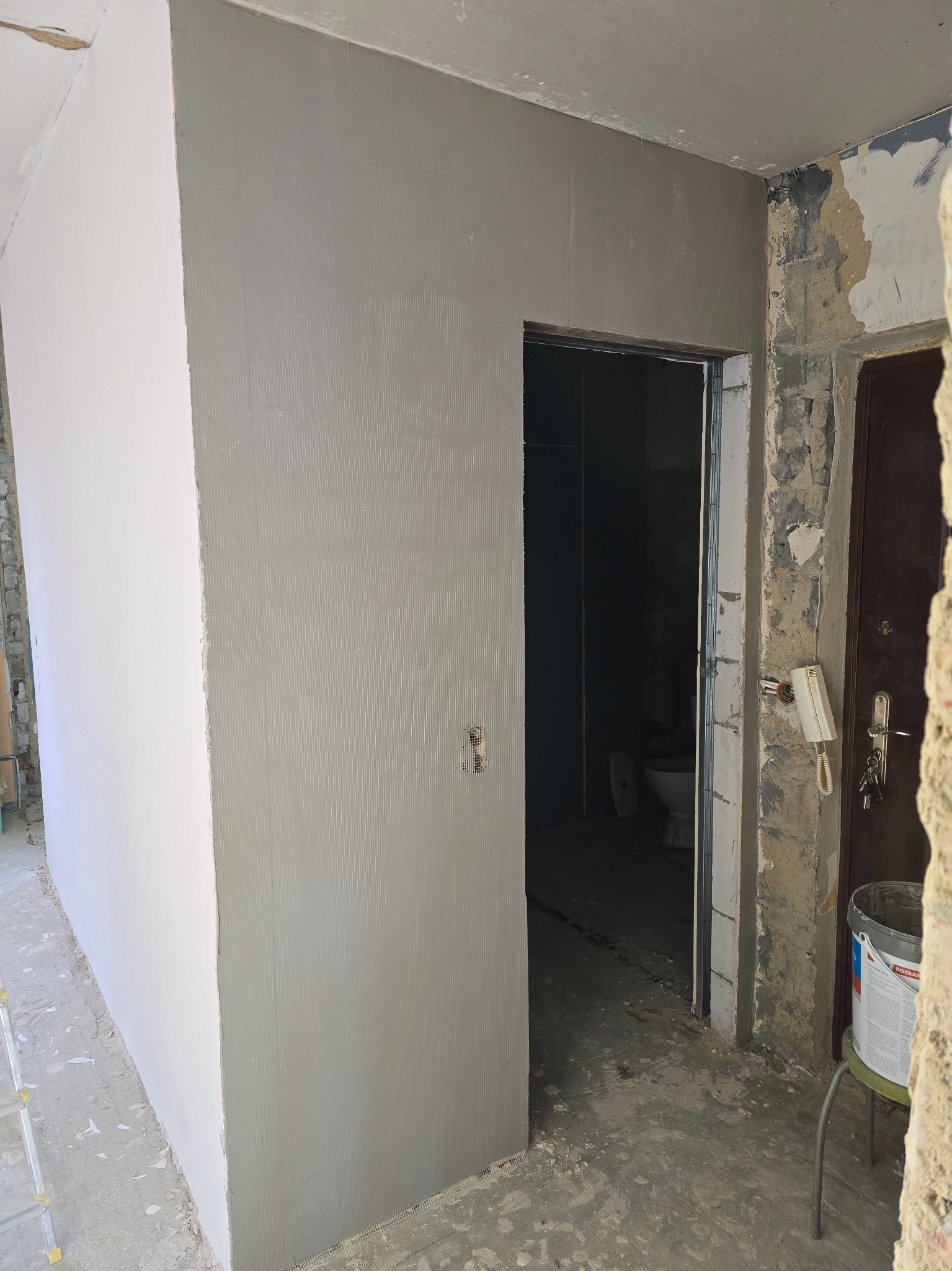 Ściana po szpachlowaniu z widoczną fakturą, fragment ściany pomalowany na biało, drzwi do łazienki w tle, widoczne ślady remontu, kubeł z farbą na stołku.