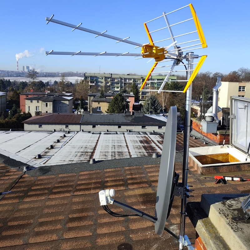 Antena satelitarna i naziemna na dachu budynku z widokiem na miasto w tle, w słoneczny dzień. Widoczne elementy montażowe i kable.