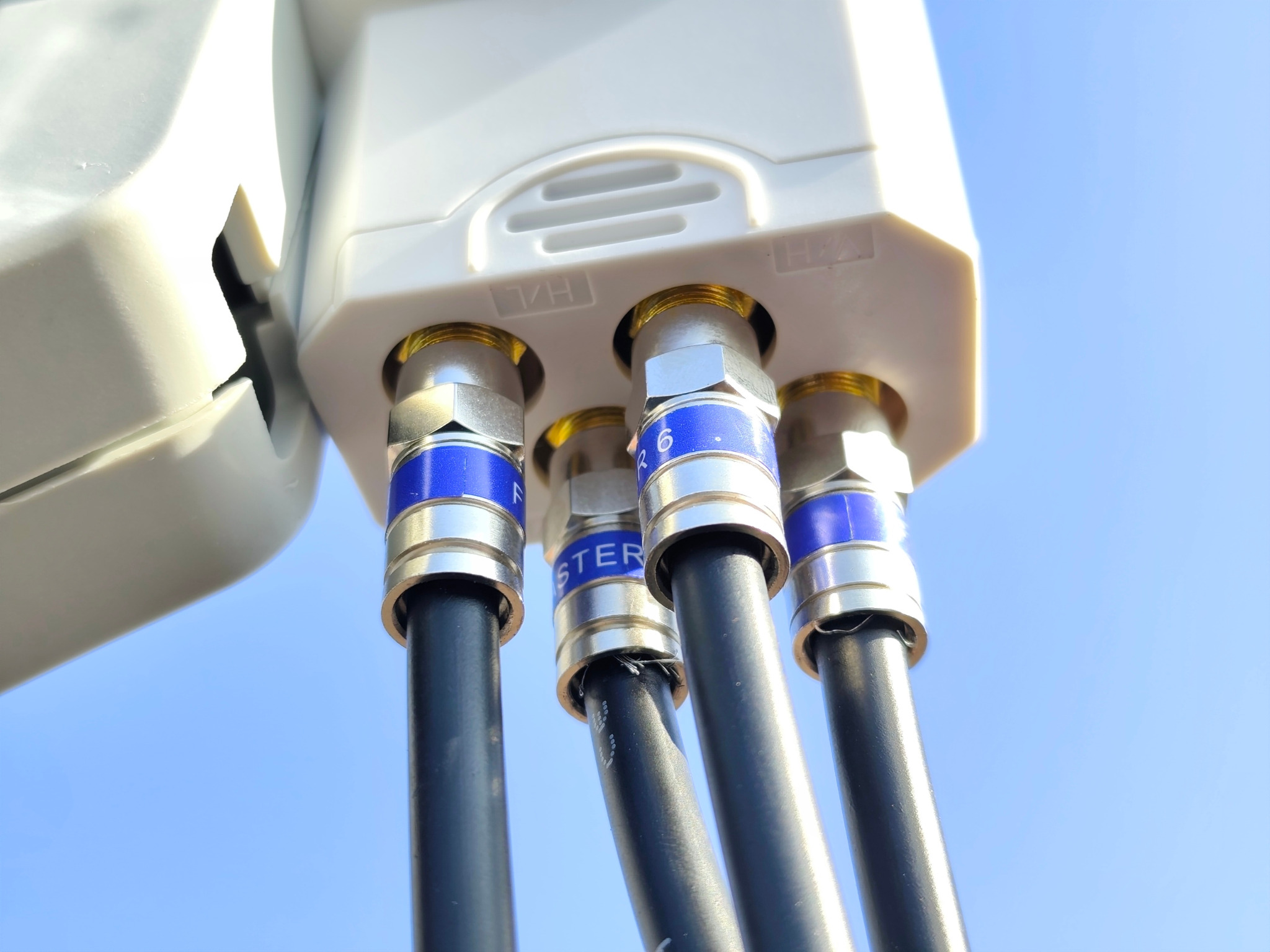 Złącza kablowe anteny satelitarnej z widocznymi kablami koncentrycznymi i niebieskimi oznaczeniami, na tle błękitnego nieba. Detal instalacji antenowej.