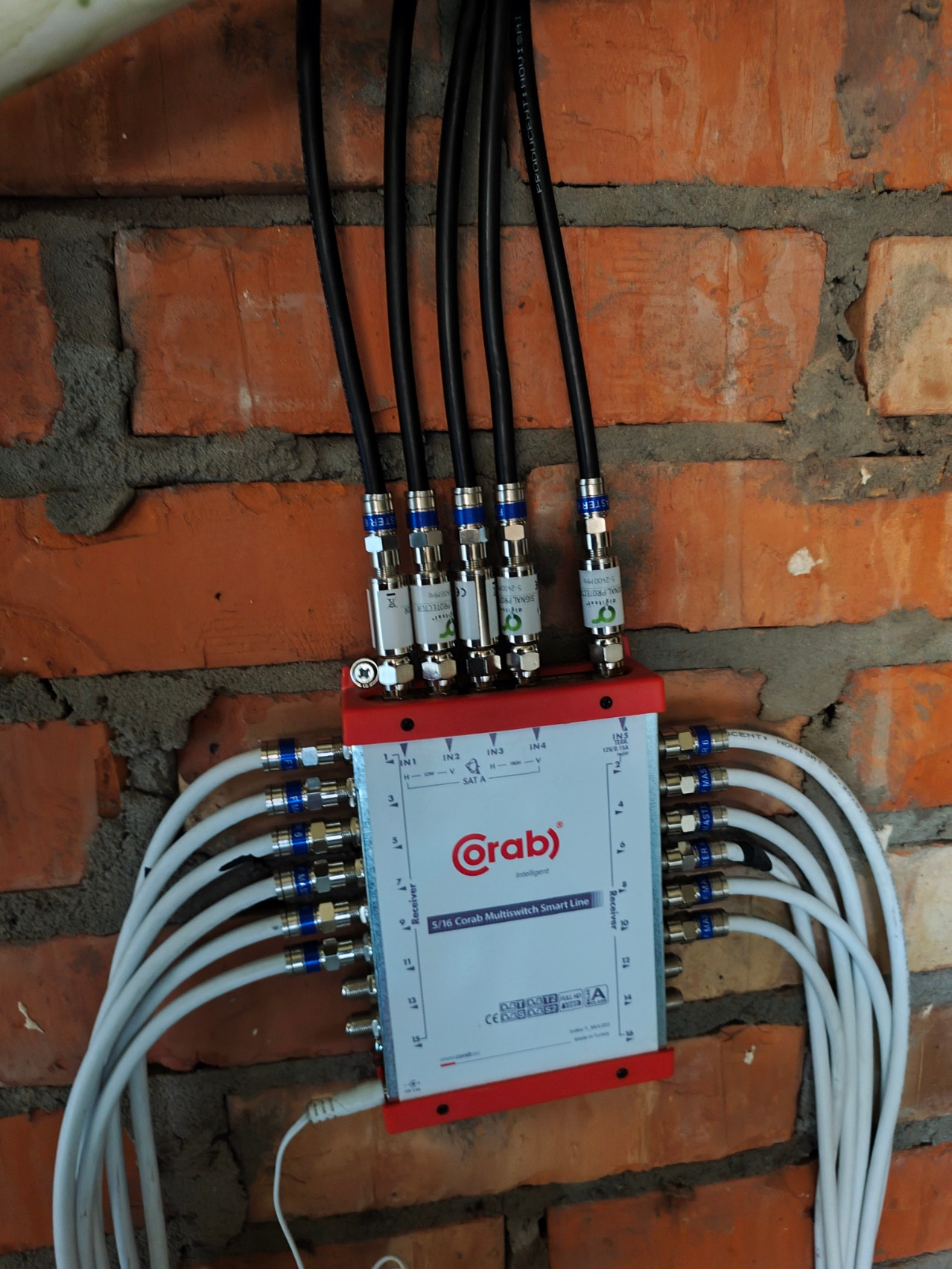 Rozdzielacz antenowy Corab 5/16 z podłączonymi kablami koncentrycznymi, zamontowany na ceglanej ścianie. Widoczne złącza i oznaczenia na urządzeniu.