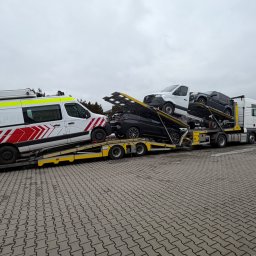 JARPOL GROUP SPÓŁKA Z OGRANICZONĄ ODPOWIEDZIALNOŚCIĄ - Wielopoziomowa laweta z trzema pojazdami, w tym ambulansem, na parkingu w Dawidach. Transport aut z zagranicy na specjalistycznej platformie. Szary dzień.