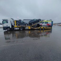 JARPOL GROUP SPÓŁKA Z OGRANICZONĄ ODPOWIEDZIALNOŚCIĄ - Ciężarówka laweta z trzema pojazdami: SUV, osobowy i ambulans. Transport aut na mokrym asfalcie, pochmurne niebo w tle. Widok z boku, lekki deszcz.