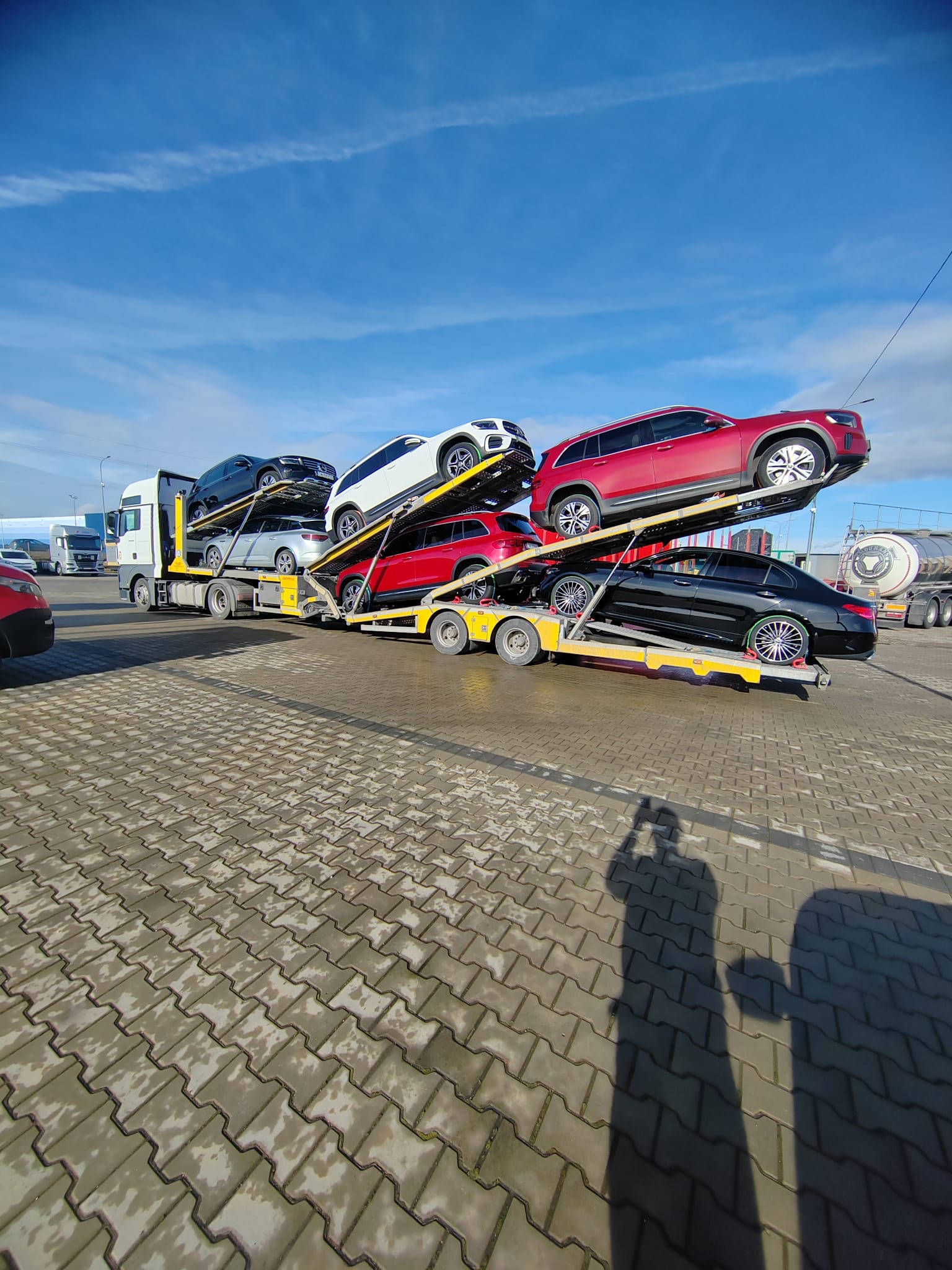 Wielopoziomowa laweta z samochodami osobowymi różnych marek i kolorów na parkingu, w tle błękitne niebo. Widoczny cień osoby robiącej zdjęcie.